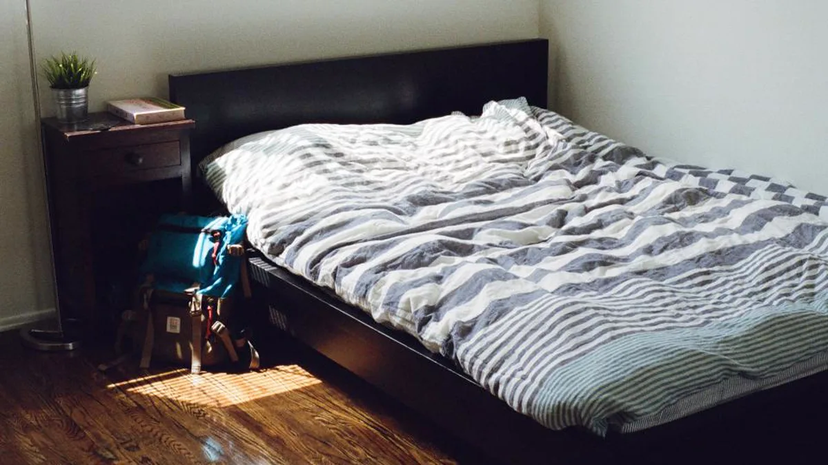 A sunlit bedroom with an unmade bed and a backpack on the wood floor
