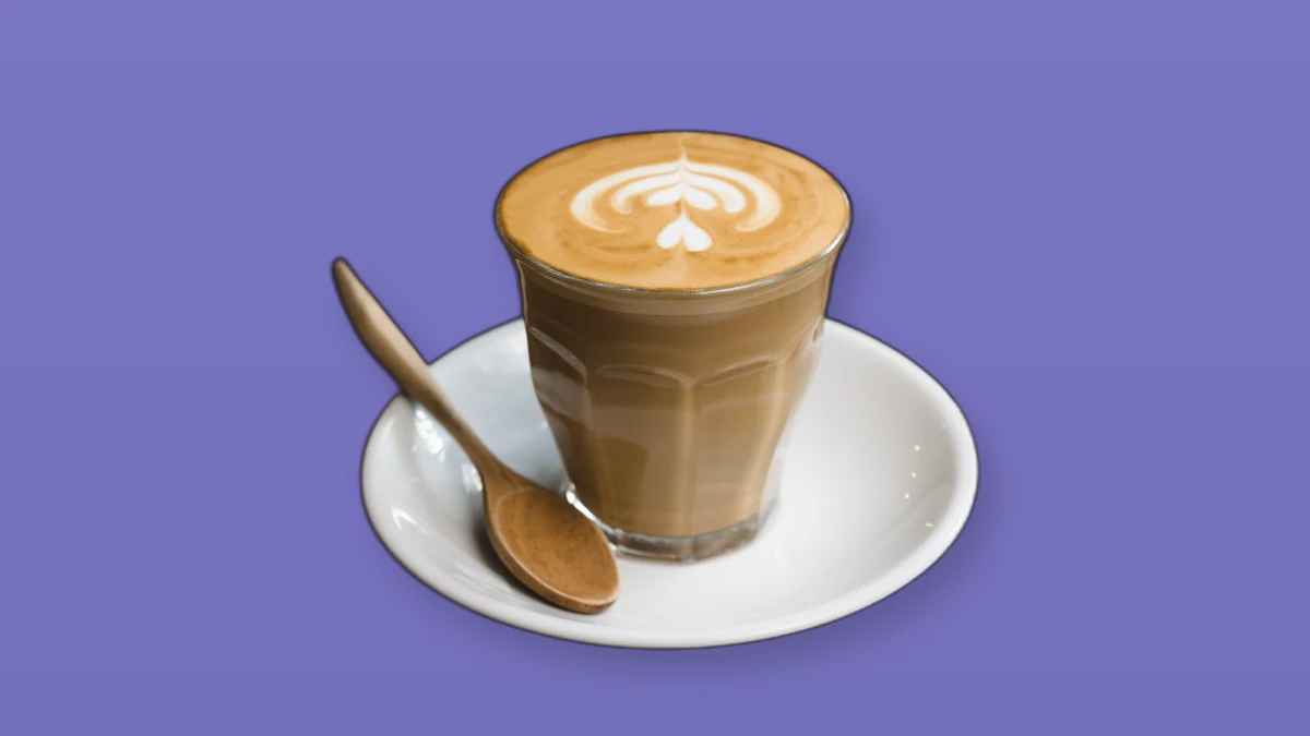 A latte in a glass on a saucer with a wooden spoon, set on a lavender background