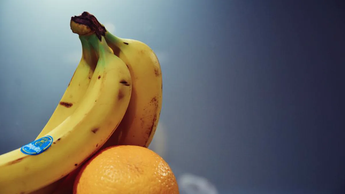 A bunch of ripe yellow bananas with brown spots resting on an orange against a blue-gray background