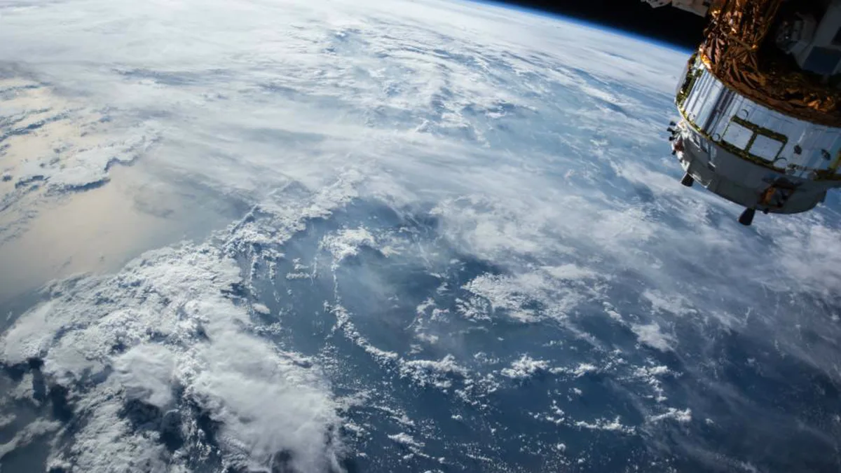 Earth seen from low orbit, with a cloud-covered ocean below and a spacecraft module at the edge of the frame