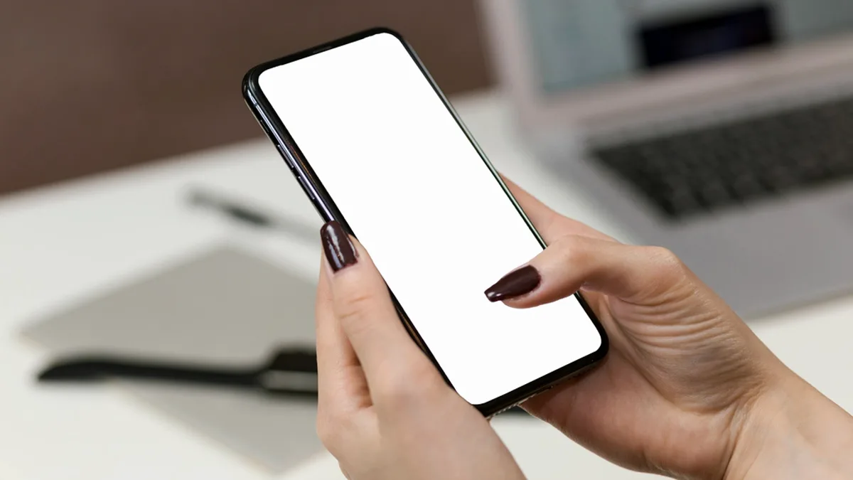 A hand holding a smartphone with a blank screen above a desk